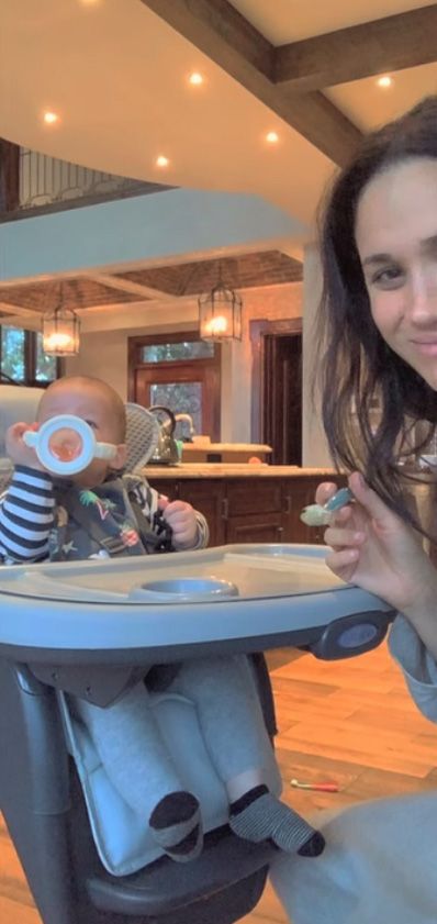 Prince Archie in a high chair drinking while Meghan Markle holds a spoon of food
