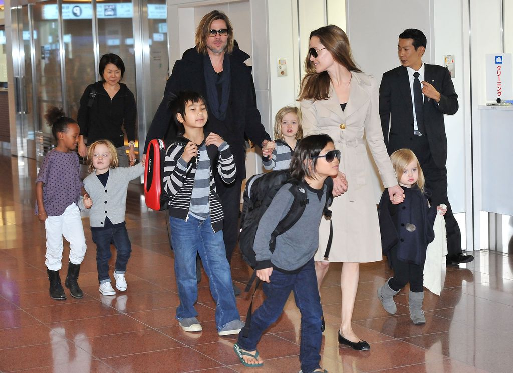 Angelina in a biege coat and sunglasses smiling at her children Maddox, Pax, Zahara, Shiloh, Knox, and Vivienne in the airport and Brad Pitt in the background