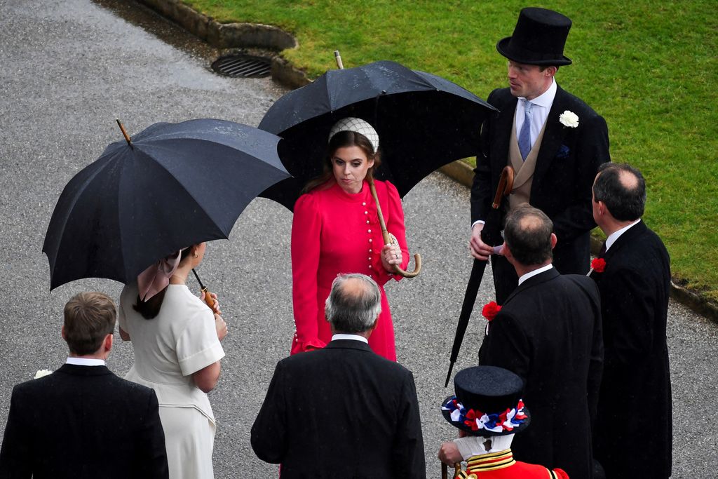 Princesa Beatrice usando vestido vermelho e bandana na festa no jardim do Palácio de Buckingham sob chuva