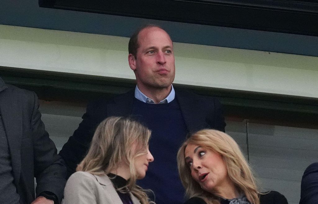 Prince William at a football match; two women are talking in front of him
