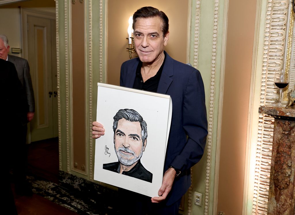George Clooney holding his portait from Sardi and smiling for camera