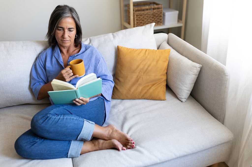 Serene mature woman enjoying a peaceful moment at home, comfortably reading a book and sipping tea on her sofa