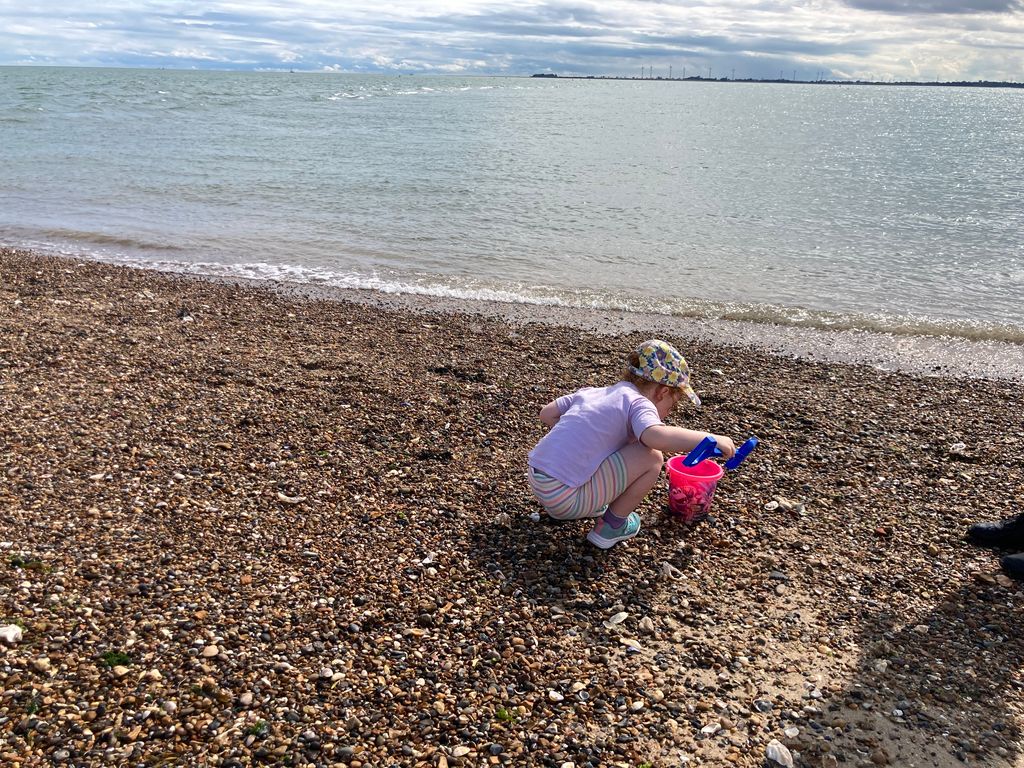 Girl wearing a hat playing on pebbly beach with a pink bucket and blue spade