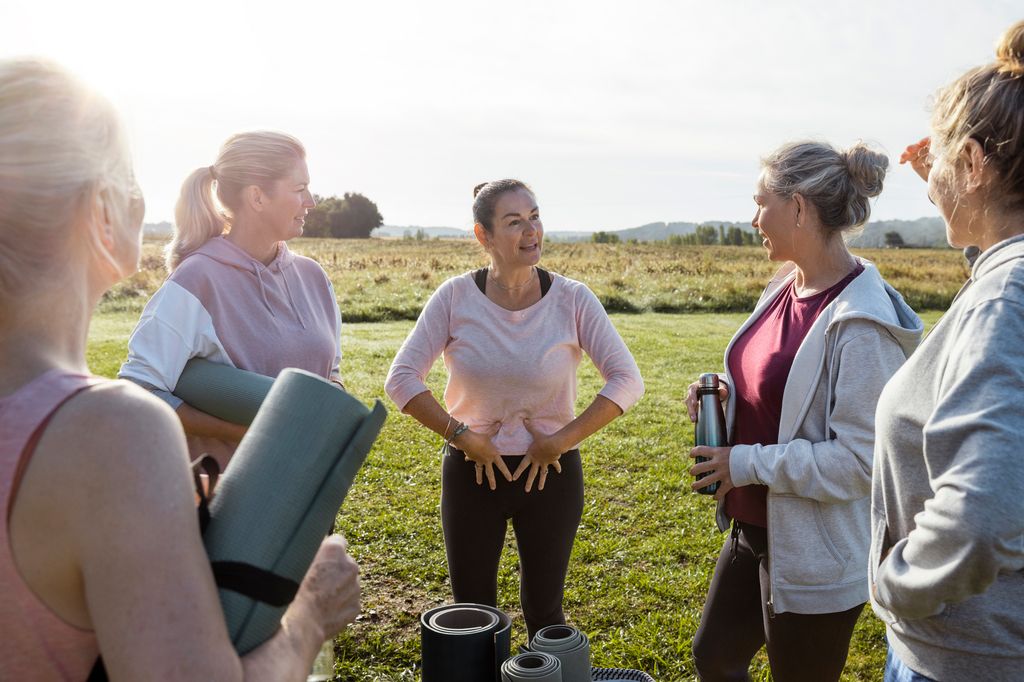 Group of mature women doing Yoga outdoors in France together while on a wellness vacation. They are all standing holding their mats ready to get started. The yoga instructor is giving advice on the exercises they will be doing.