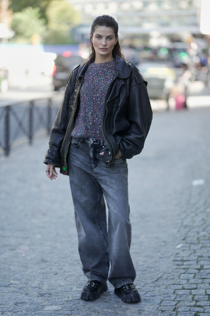 Isabeli Fontana is seen wearing a multicolored knit sweater under a black oversized leather jacket, paired with faded wide-leg grey jeans during the Womenswear Spring Summer 2026 as part of Paris Fashion Week 