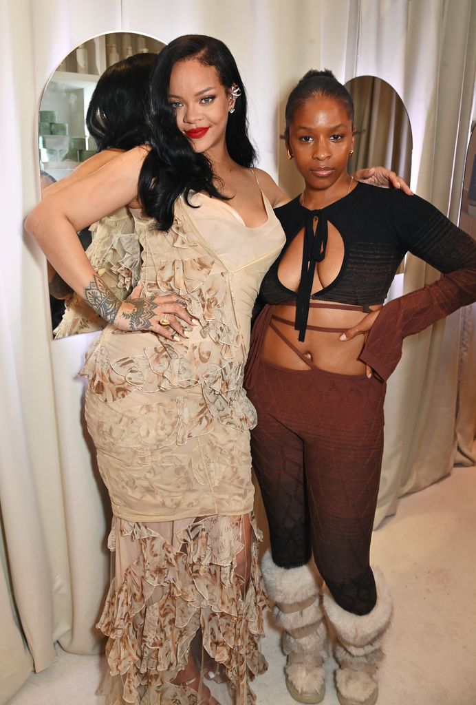 Rihanna stands beside Julie Adenuga, both dressed in creative, textured outfits. Rihanna's nude-toned, floral-inspired dress contrasts with the darker, unique attire of her companion.