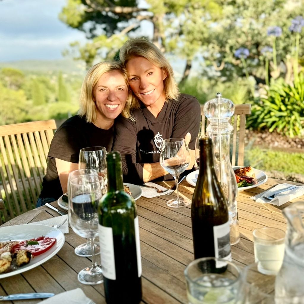 Two women smiling outside in the sun drinking wine 