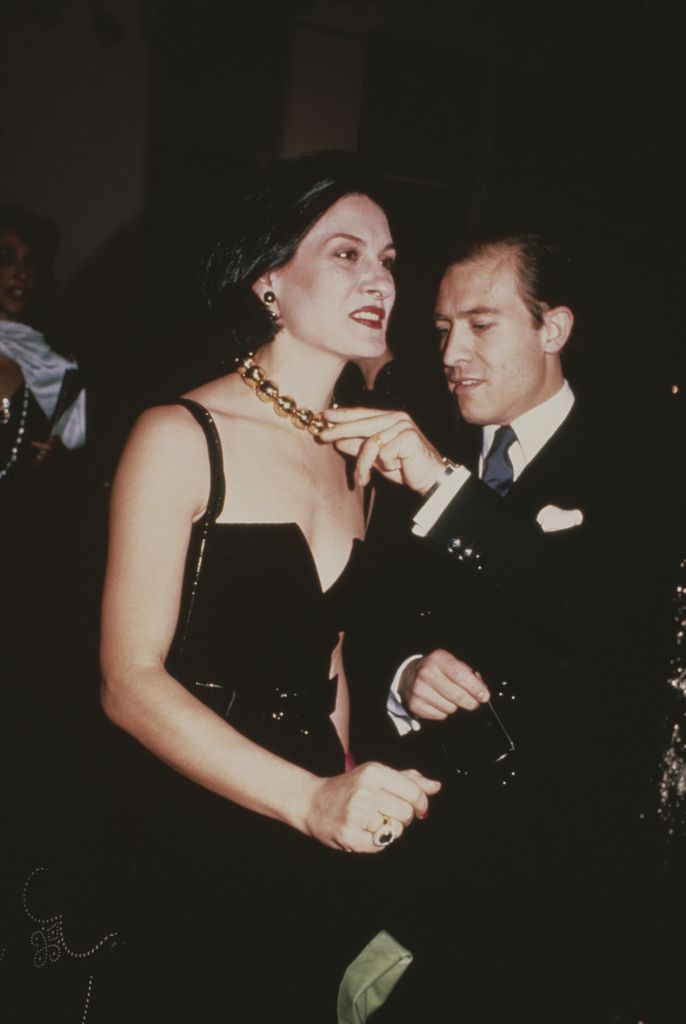 Paloma Picasso wears a minimalist black gown with a bold chunky gold chain necklace at the Met Gala, exuding a powerful and pared-down glamour with slick hair and sharp lipstick.