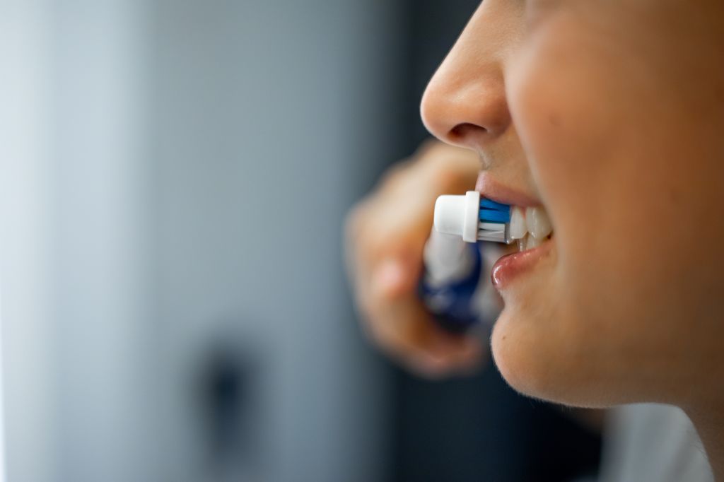 Close-up of up mouth of a young person holding electric toothbrush in hand, smiling and brushing white teeth with modern equipment