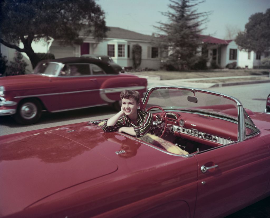 American actress and singer Debbie Reynolds smiles while sitting in the driver's seat of a red convertible Ford Thunderbird, turning in her seat to rest her right elbow on the rear of the car,