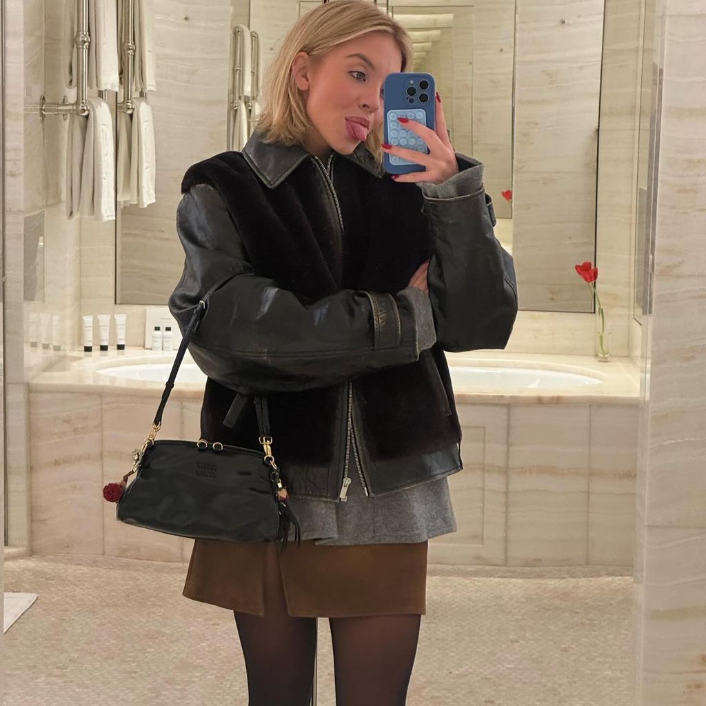 Sydney Sweeney takes a selfie in the miror wearing a black jacket, brown mini skirt and black handbag and playfully pokes out her tongue