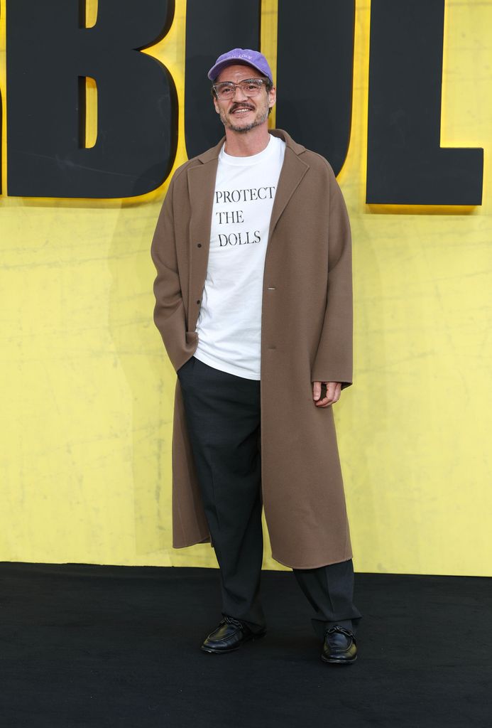 Pedro Pascal stands confidently against a bright yellow backdrop with the word "UNBEARABLE" in massive black letters. He wears a long, unbuttoned camel wool coat that flows to mid-calf, a crisp white T-shirt printed with the bold slogan "PROTECT THE DOLLS," wide charcoal trousers, and black leather dress shoes. A lavender baseball cap rests atop his head, and he finishes the look with clear-rimmed glasses and a trimmed mustache and beard. He smiles warmly, one hand in his coat pocket, relaxed and self-assured.