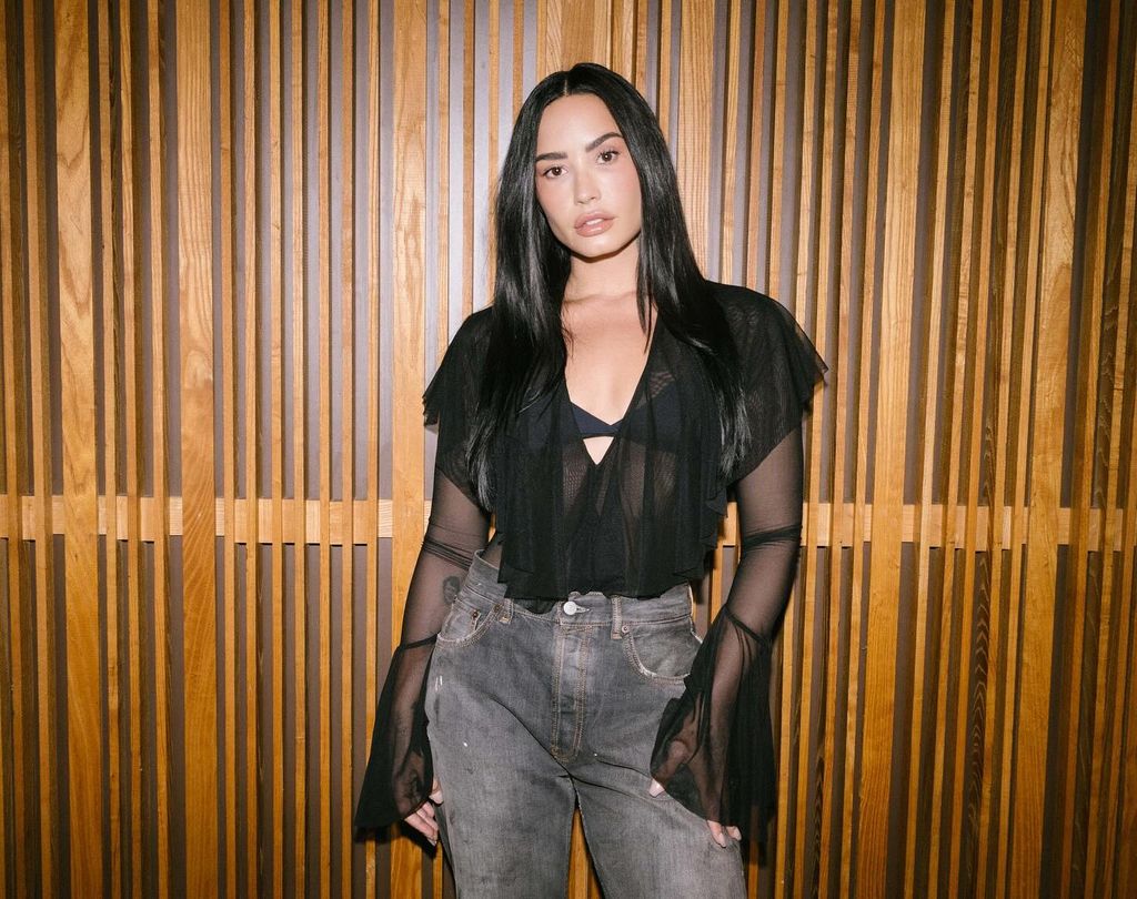 photo of demi lovato in jeans and plunging black top posing in front of wood wall