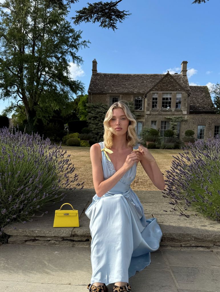 Elsa Hosk est assise dans un jardin de campagne, une grande maison est derrière elle. Elle ajuste la sangle sur sa robe bleu ciel et jaune, son sac à main jaune perché à côté d'elle