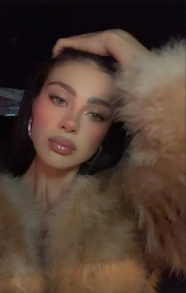 Nicola Peltz Beckham wears a pale furry coat and raises her hand above her head, combing through her long dark hair. She is also wearing encrusted hoop earrings, glitter eyeshadow and lipgloss as she poses in her car