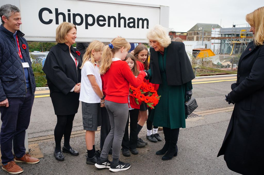 Queen Camilla being presented with a crocheted poppy wreath by group of children on tarin platform