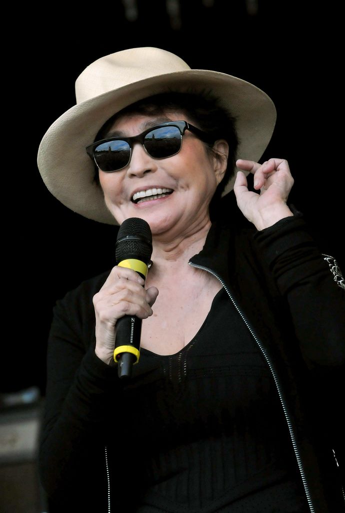 Yoko Ono tient son doigt gauche pincé près de son oreille tandis que sa main droite tient un microphone. Le chanteur porte un large chapeau crème et des lunettes de soleil.