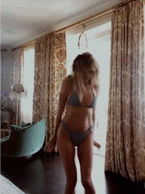heidi stands in a bikini in a bedroom with two huge floor to ceiling windows and luxurious floral curtains are draped around them falling to a dark hardwood floor