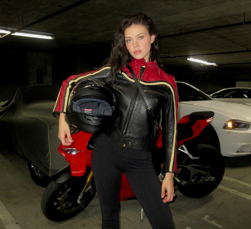 Nicola Pelta Beckham poses in front of her motorbike in a red, beige and black leather jacket and holds a helmet under her right arm