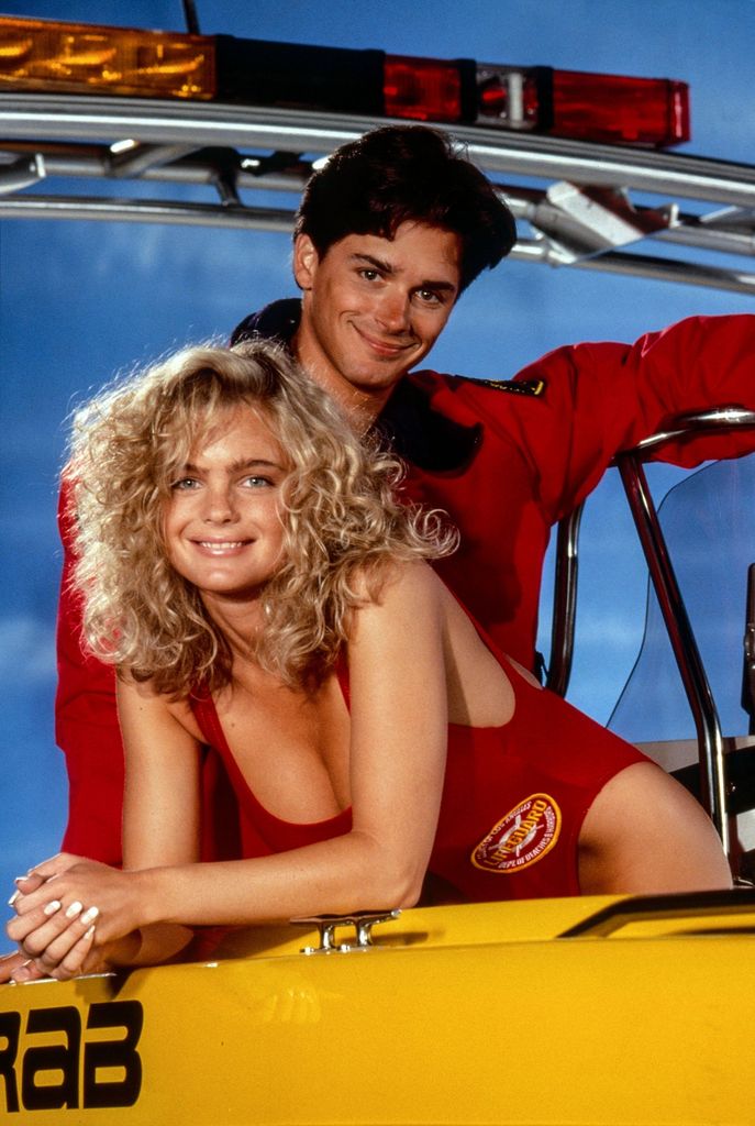 Erika leaning over a lifeguard boat smiling in a swimsuit with Baywatch co-star
