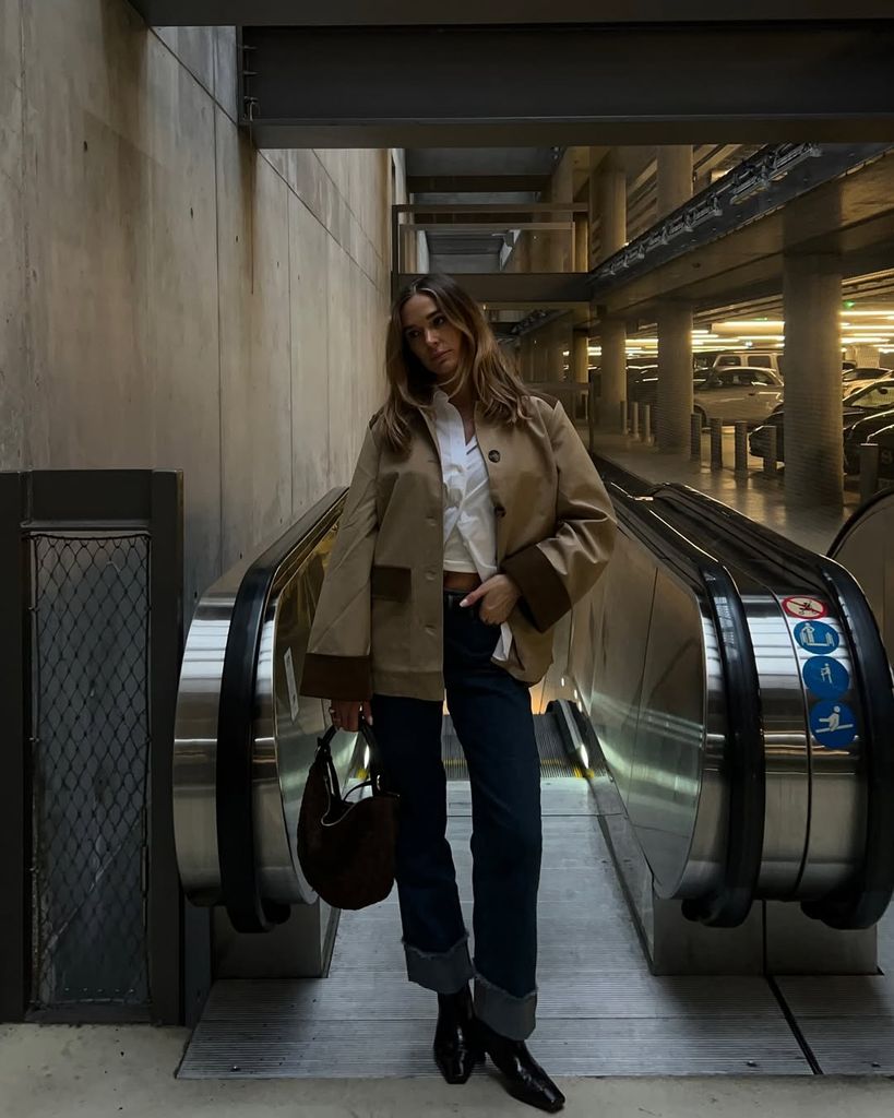 Scandi influencer Tina Maria stuns in a cropped trench jacket, cuffed denim and heeled boots. A classic white shirt peeks out beneath the coat as she poses at the top of an escalator.