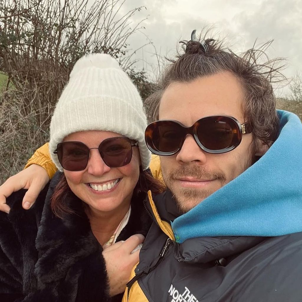 Harry Styles wears a North Face jacket and sunglasses as he puts his arm around his mother, Anne Twist. Anne is smiling and wears a black fluffy velvet coat, sunglasses and a white hat.