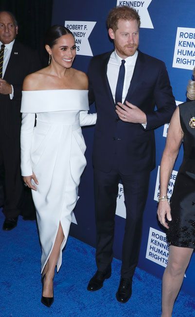 meghan wears a white off the shoulder dress as she stands with prince harry wearing a blue suit on a blue carpet