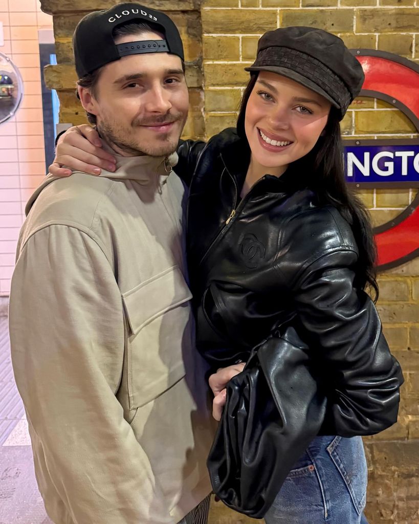 Nicola Peltz and Brooklyn Beckham pose together at a London Underground station, standing in front of a brick wall with a roundel sign partially visible behind them. Brooklyn, wearing a beige jacket and a black baseball cap with "CLOUD 23" embroidered on it, has a slight smile and light stubble. Nicola, smiling brightly, wears a black leather jacket with a Chanel logo, a brown newsboy cap, and blue jeans. Her arm is wrapped around his shoulder, and they appear happy and relaxed.