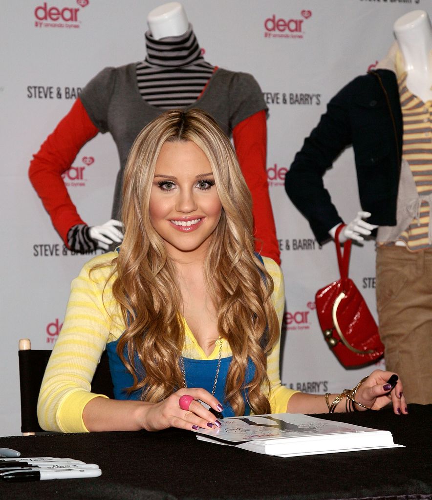 Amanda Bynes est assise derrière un bureau avec une pile de pages d'autographes pour le lancement de sa ligne de vêtements, dont des échantillons sont posés sur des mannequins derrière elle.