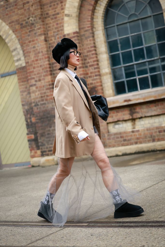 Charlene Davies wearing black fur hat, beige blazer, Havaianas thongs in pockets, Prada necktie, black clutch, white sheer pants during Australian Fashion Week 2025 on May 15, 2025 in Sydney, Australia