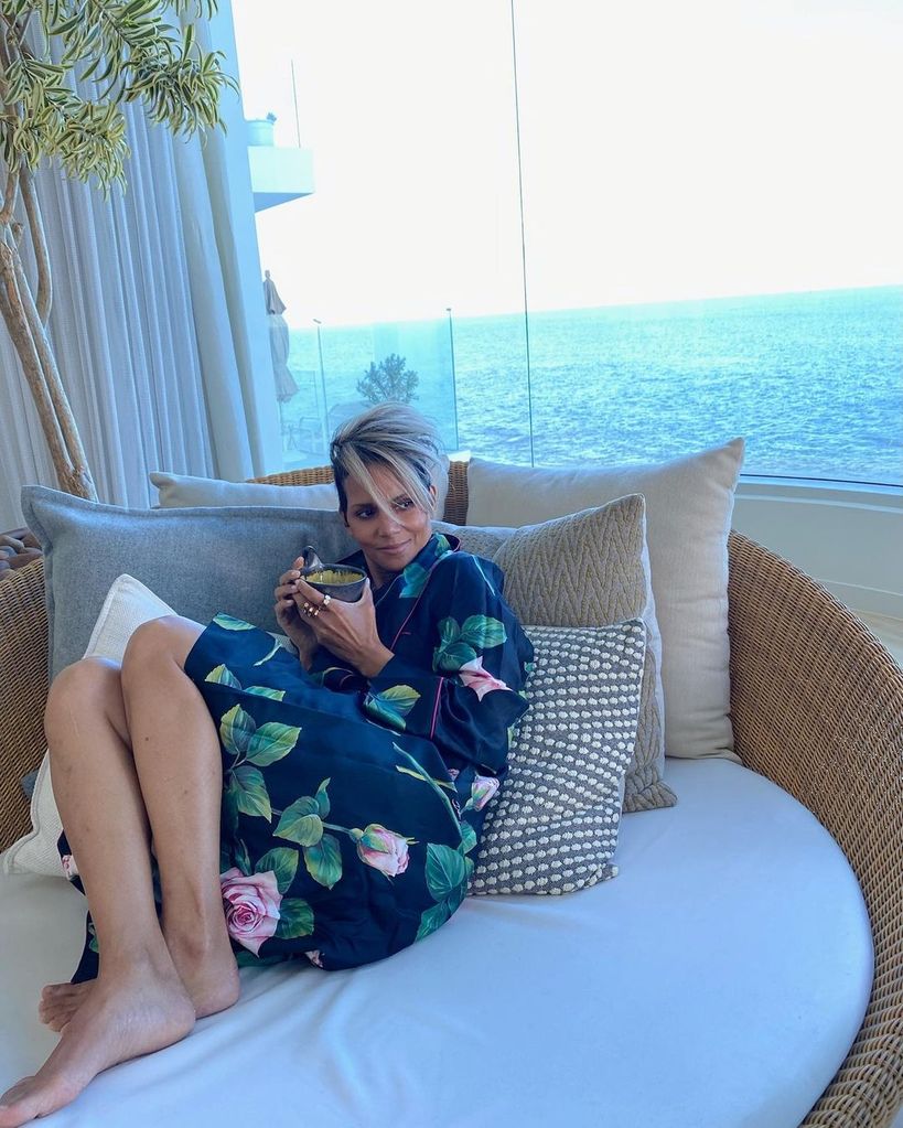 Halle Berry reclining on a round couch holding a mug and smiling, the sea stretches out behind her in the view from her window