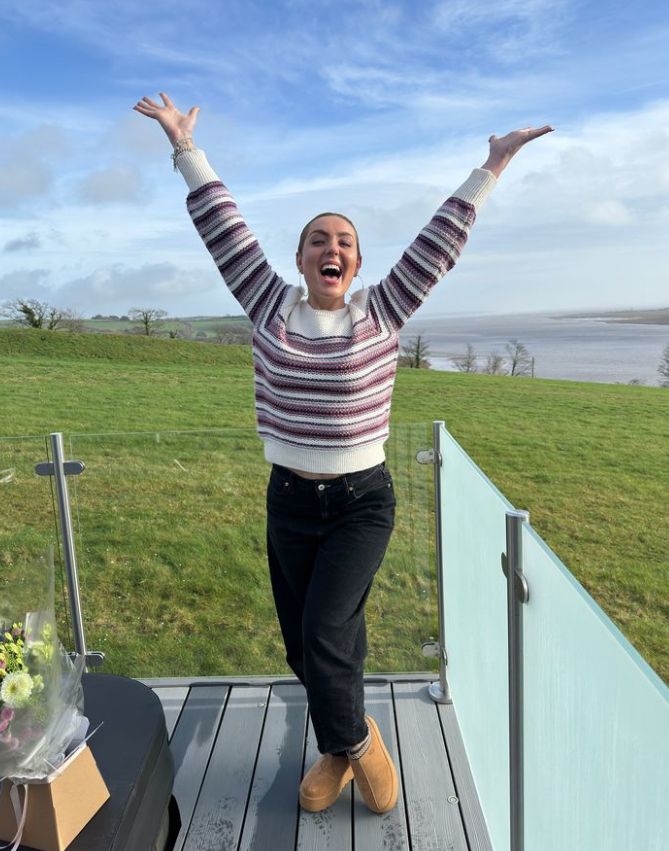 Amy Dowden on a balcony in front of grassy field with her hands in the air