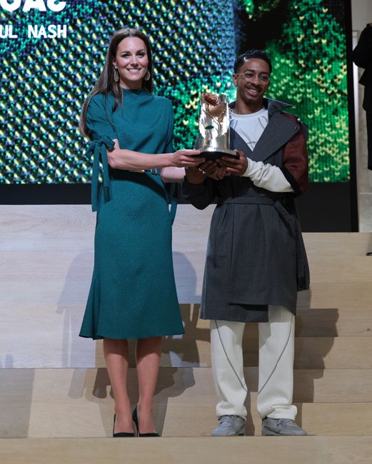 kate holds a trophy up with a young man on a stage and her dress is a dark teal hue which stops below the knee and elbow with bows on the sleeve