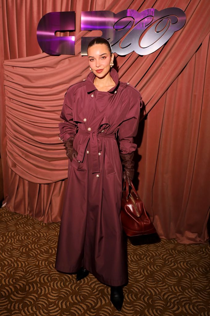 Jackie Apostel celebrates the launch of Cultedxo and poses in a deep red trench coat with matching leather gloves and bag. Her hair is slicked back into a bun and she is wearing bright red lipstick.