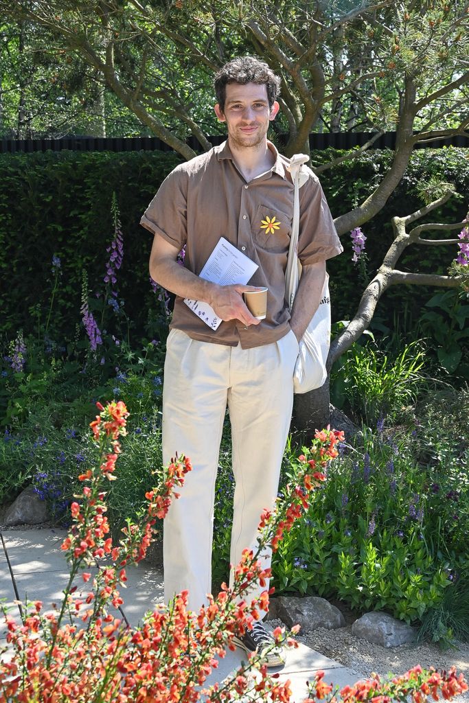 Josh O'Connor attends the Stroke Association's Garden for Recovery at RHS Chelsea Flower Show  in a brown button up shirt and beige trousers