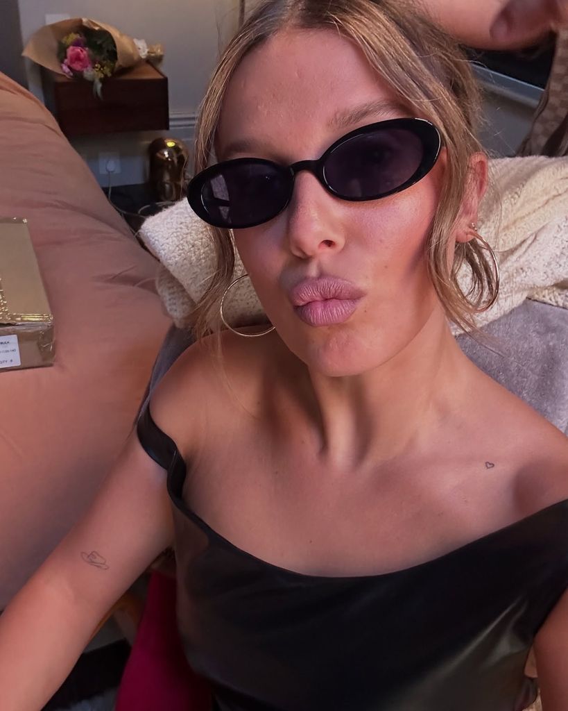 MIn this close-up selfie, Millie Bobby Brown is seated indoors, facing the camera with a pouty expression. She is wearing sleek, oval-shaped black sunglasses reminiscent of early 2000s fashion and a one-shoulder black leather-look top, which reveals minimalistic tattoos—a small outline of a heart near her collarbone and the Vivienne Westwood orb on her forearm. Her light brown hair is styled up with a few soft tendrils framing her face. Gold hoop earrings complete the Y2K-inspired look. The background includes a pink bedspread, a bouquet of flowers on a nightstand, and a gold-wrapped gift box, suggesting a celebratory or intimate setting.