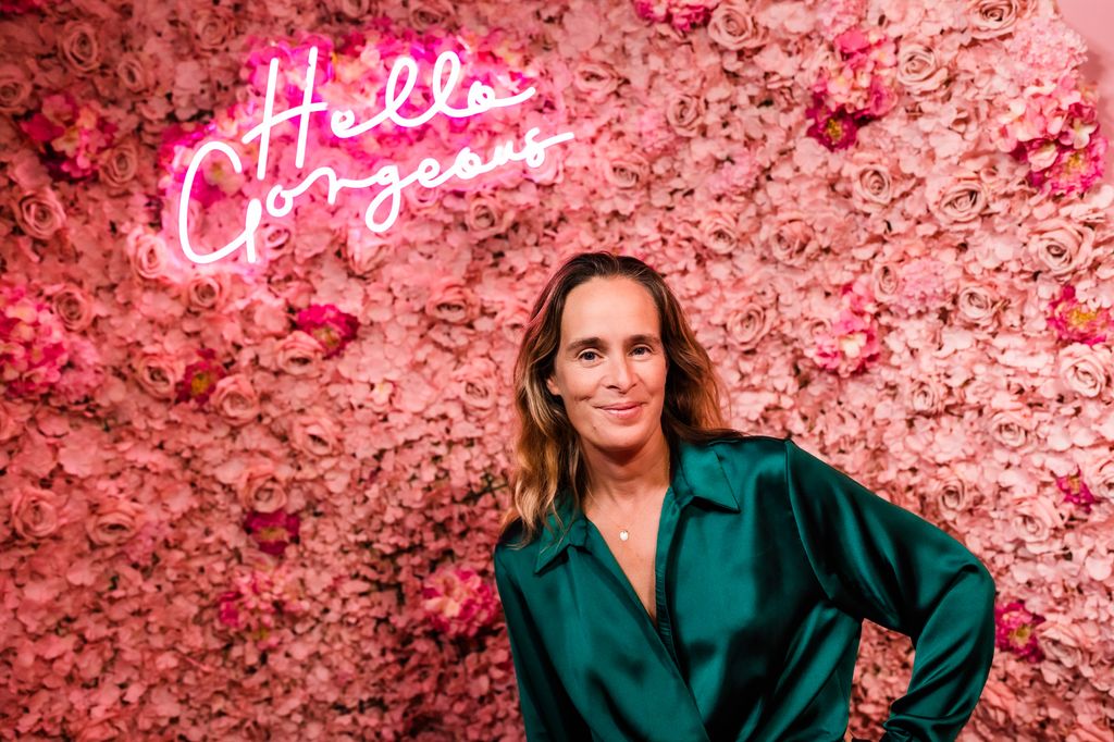 woman in a silky green shirt posing in front of a pink flower wall with hello gorgeous neon light