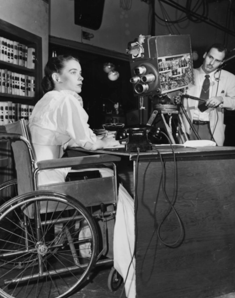 Actress Susan Peters sitting behind a desk in her wheelchair, in front of a WPTZ television camera filming her show 'Miss Susan', circa 1951