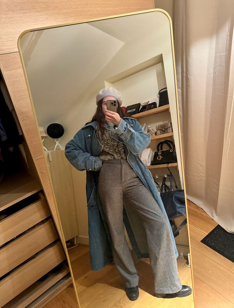 Natalie Salmon poses in a mirror selfie wearing Sezane trousers paired with a frilly blouse, a long denim coat, black boots, and a white beret, blending Parisian chic with vintage flair in a cozy setting.