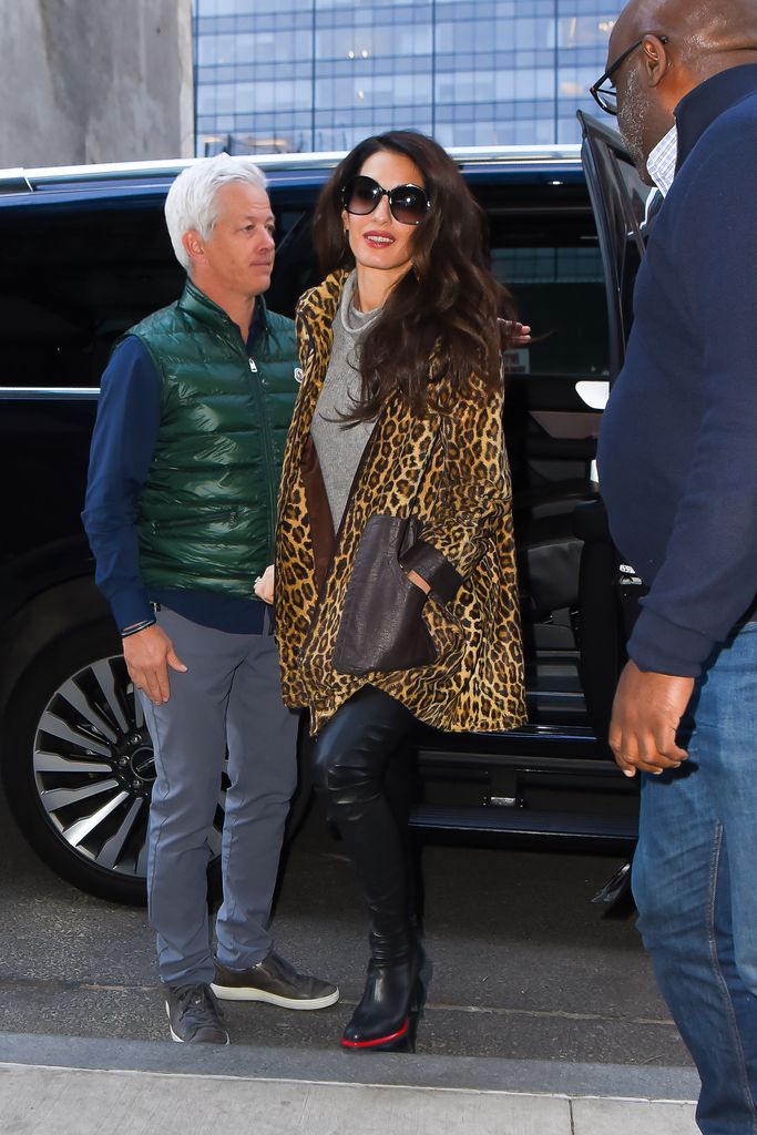 Amal Clooney walks down the street in black thigh high boots, a grey sweater mini dress and a leopard print coat. She wears sunglasses and carries a black shoulder bag. 