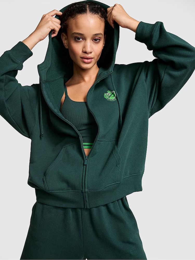 A model showcases a Victoria's Secret PINK green hoodie paired with matching sweatpants. She pulls the hood up, exuding sporty and casual vibes.