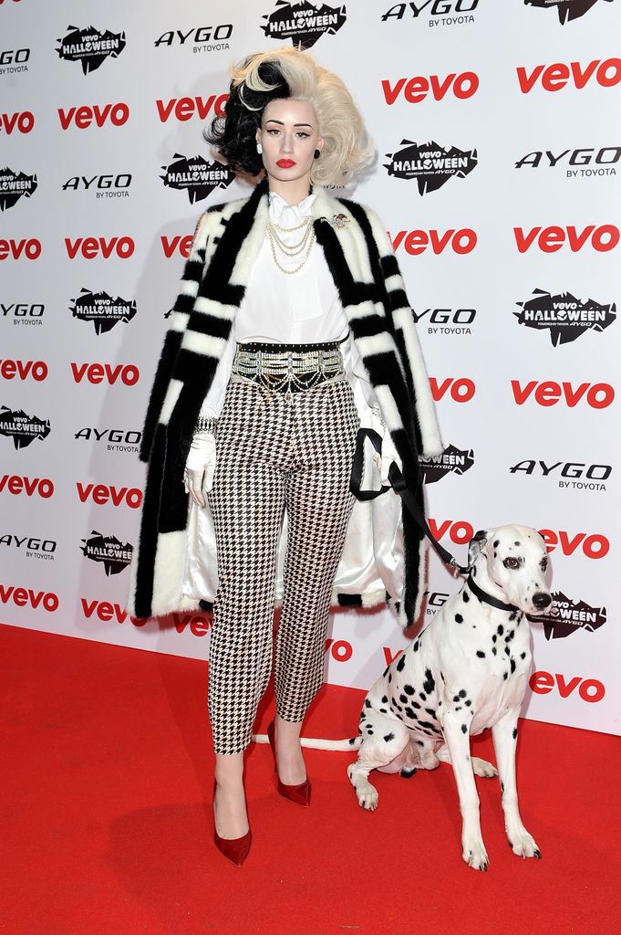 Iggy Azalea dressed as Cruella de Vil in a black-and-white fur coat, houndstooth pants, and half-black, half-white hair. They stand on a red carpet with a Dalmatian dog by their side.