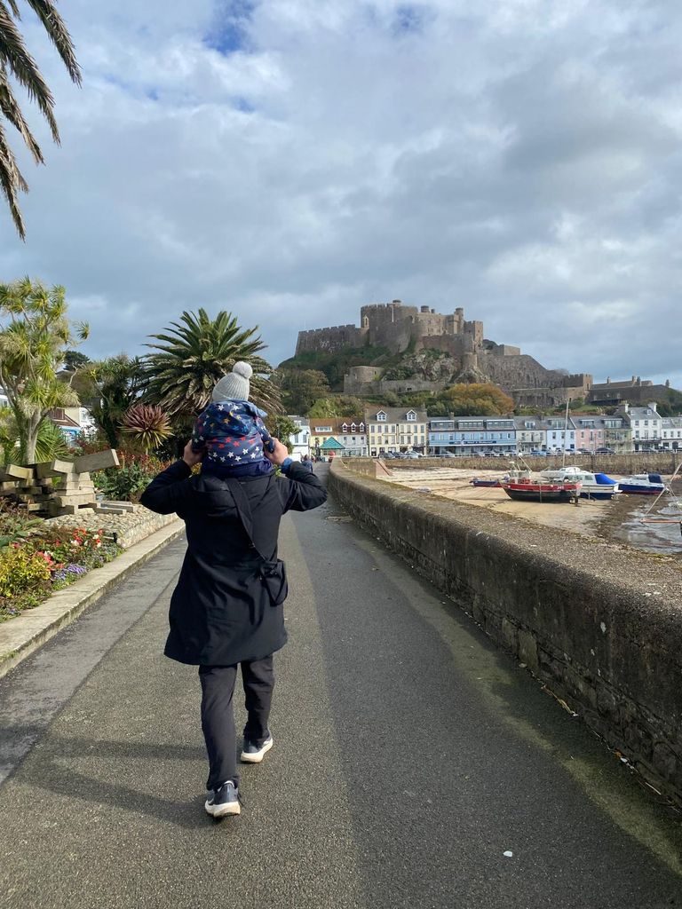 Man with boy on his shoulders walking along a path with a harbour and castle in the background
