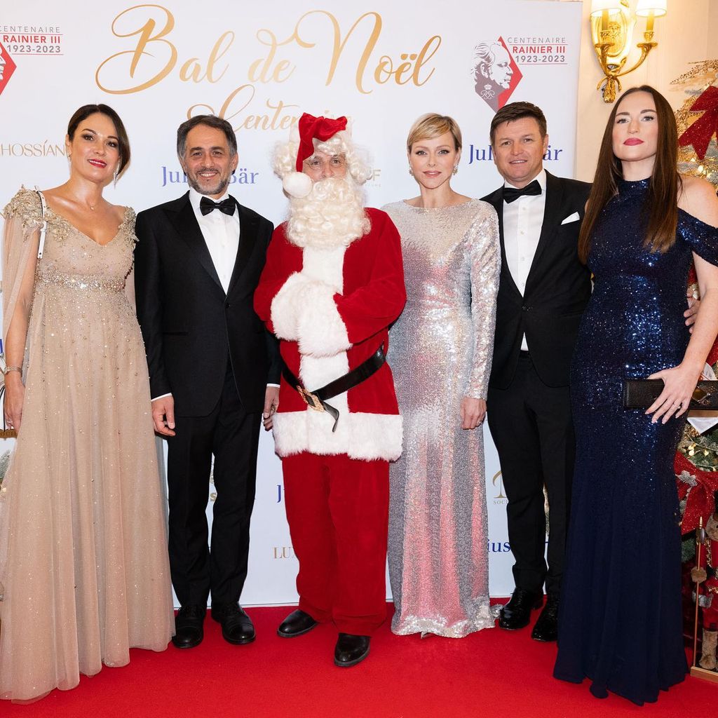 Princess Charlene wears a silver dress and stands next to someone wearing a Santa costume