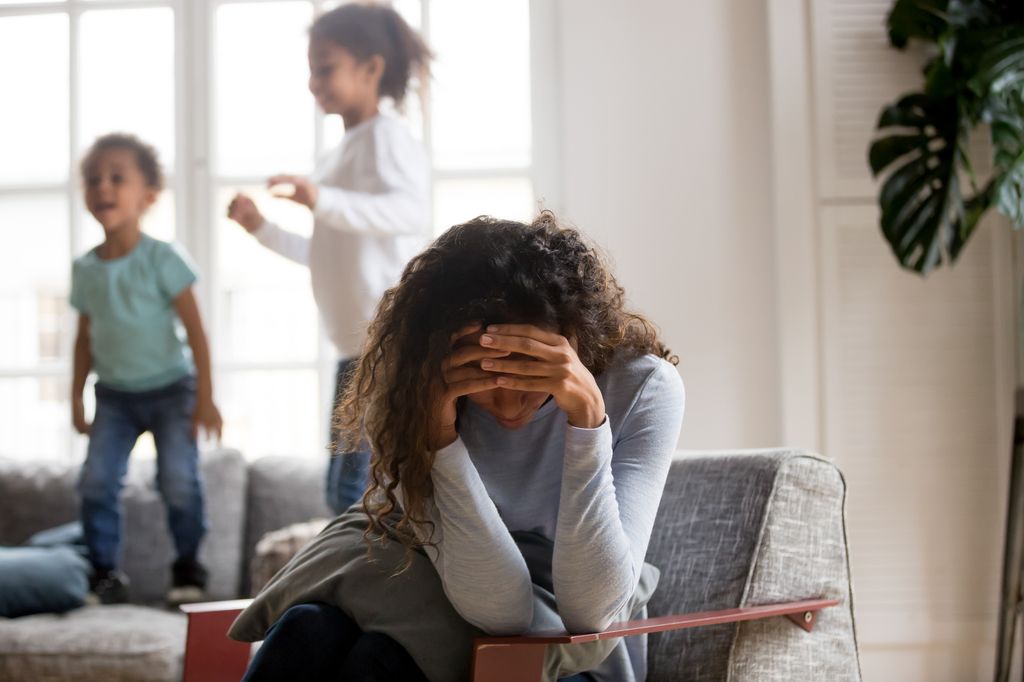 Woman holding her head with hands with playful kids jumping on couch in the background