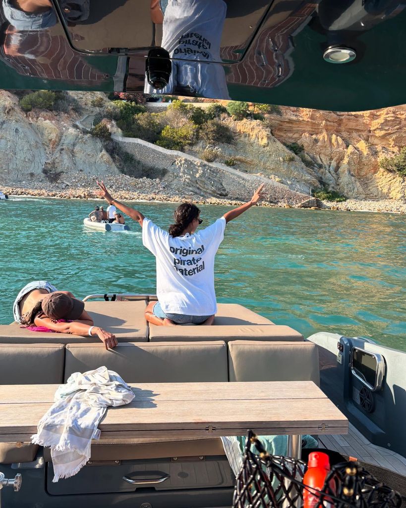 Maya Jama sits on a luxury yacht, hands in the air, wearing an oversized whit t-shirt emblazoned with 'Original Pirate Material'