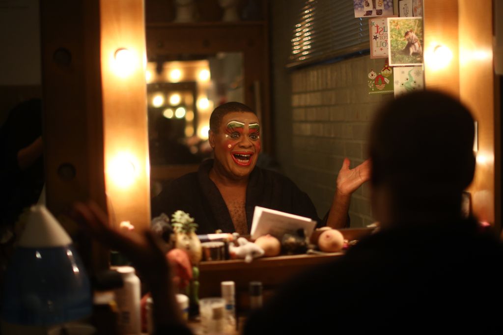 A photo of Clive Rowe looking in the mirror wearing colourful makeup