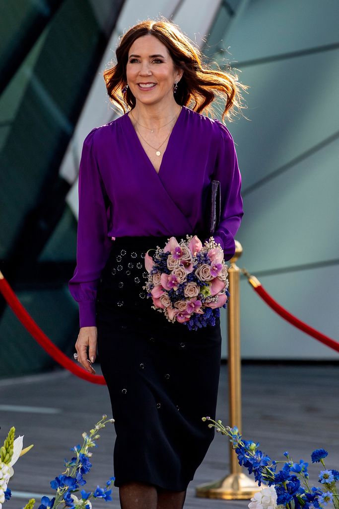 Queen Mary in a pencil skirt and purple top holding flowers