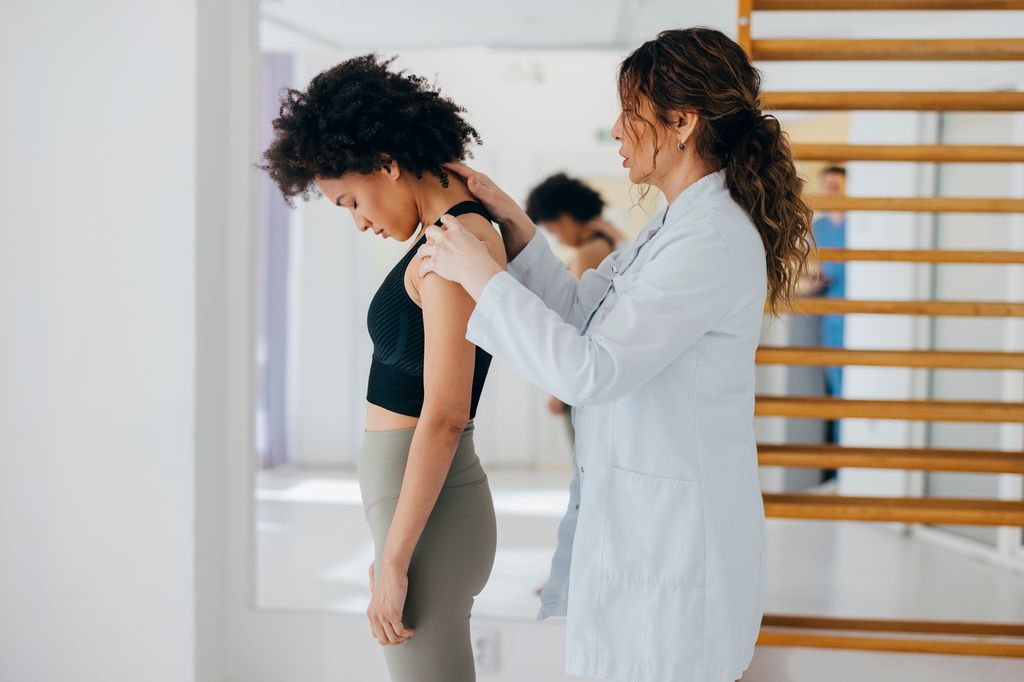A medical professional examining a patient's neck and shoulder alignment in a bright clinical setting with a wooden wall ladder