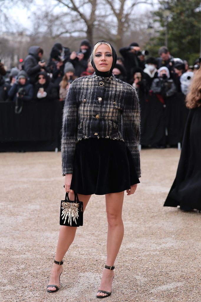 Pixie Lott wore a cropped plaid tweed jacket with golden buttons, a black velvet mini skirt, a celestial-themed handbag, strappy heels, and a chic headscarf. Parisian winter elegance!